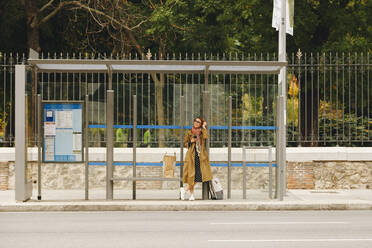 Woman in trench coat waiting at bus stop - MRRF01652