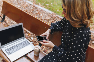 Businesswoman using mobile phone on bench at park - MRRF01659