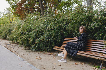 Lächelnde Frau sitzt auf einer Bank in einem öffentlichen Park - MRRF01670
