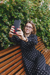 Smiling woman taking selfie through smart phone on bench - MRRF01671