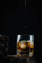 Whiskey drops falling on ice cubes served in crystal glass placed on rough surface against black background - ADSF31397