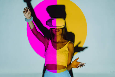 Surprised female with raised arm exploring virtual reality in headset while standing in pink and yellow projector light on gray background - ADSF31398