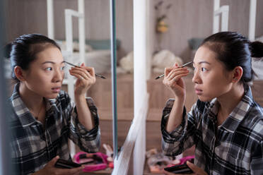 Focused charming ethnic female applying eyebrow pencil while doing makeup and looking in mirror - ADSF31403