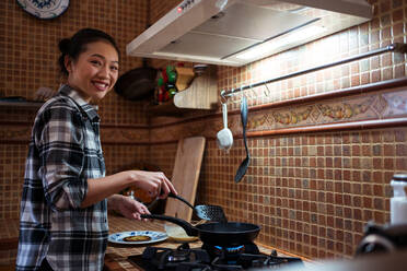 Seitenansicht einer jungen glücklichen Asiatin in Freizeitkleidung, die leckere Pfannkuchen mit einem Spatel zubereitet, während sie in der Küche steht und in die Kamera schaut - ADSF31408