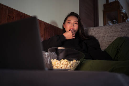 Young Asian female wearing casual clothes sitting on couch and eating popcorn at home watching movie on laptop - ADSF31409