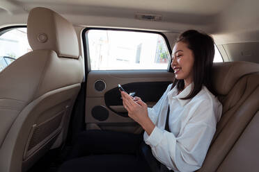 Side view of asian female passenger in formal clothes browsing cellphone while sitting on backseat in taxi and looking away with interest - ADSF31414