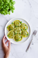 Top view of crop anonymous person at marble table with raviolis near fork and fresh basil leaves in light kitchen - ADSF31425