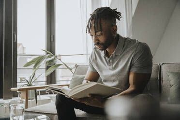 Young bearded man reading book on sofa - MFF08298