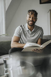 Smiling man holding book on sofa at home - MFF08299