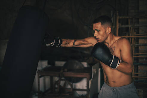 Hemdloser junger Sportler übt Boxen mit Boxsack im Fitnessstudio zu Hause - ACPF01401
