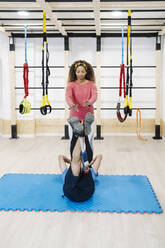 Weiblicher Fitnesstrainer, der einem Mann mit Behinderung im Fitnessstudio hilft - JCZF00861
