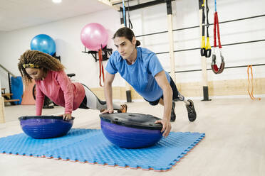 Behinderter Mann mit Freund beim Training auf dem Balance-Ball im Fitnessstudio - JCZF00863