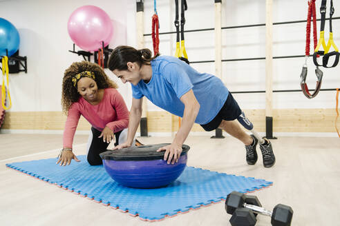 Lächelnde Fitness-Trainerin, die einem behinderten Mann bei seinen Übungen mit dem Balance-Ball im Fitnessstudio hilft - JCZF00864