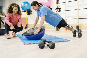 Lächelnde Lehrerin, die einem behinderten Mann bei Liegestützen auf einem Balanceball im Fitnessstudio hilft - JCZF00865