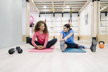 Smiling woman doing stretching exercise with disabled friend at gym - JCZF00872