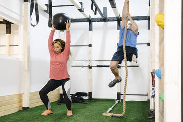 Behinderter Mann klettert an einem Seil und balanciert einen Medizinball im Fitnessstudio - JCZF00876