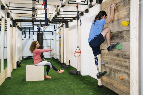 Mann mit Behinderung klettert in der Nähe eines Freundes in einem Fitnessstudio an der Wand - JCZF00878