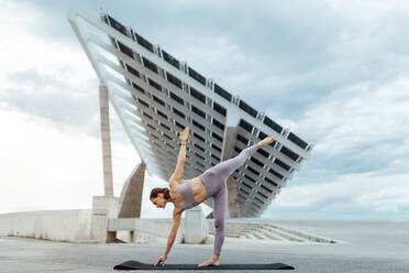 Aktive Frau in Sportkleidung macht Yoga-Asana auf einer Matte, während sie auf der Straße in der Nähe eines Solarpanels in Barcelona steht - ADSF31427