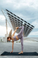 Full body of active woman in sportswear doing yoga asana on mat while standing on street near solar panel in Barcelona - ADSF31428