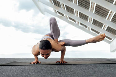 Ganzer Körper einer kräftigen, barfuß gehenden Frau in Sportkleidung beim Üben der Maksikanagasana-Haltung auf der Straße in der Nähe einer Photovoltaikanlage während eines Yoga-Trainings in der Stadt - ADSF31432