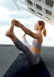 Ganzkörper-Seitenansicht einer sportlichen Frau in Activewear beim Üben der Ubhaya Padangushthasana-Haltung in der Nähe einer modernen Photovoltaikanlage während eines Yoga-Trainings auf der Straße - ADSF31435