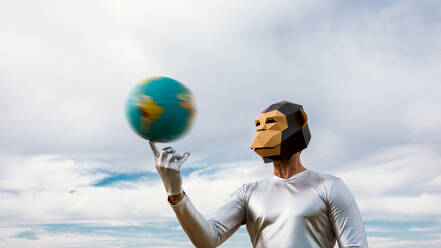 Unerkennbare Person mit geometrischer Affenmaske, die einen Ball in Form der Erde vor einem bewölkten blauen Himmel dreht - ADSF31441