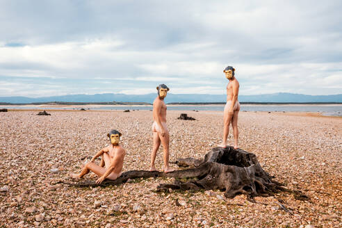 Nackte Männer mit Affenmasken, die in die Kamera schauen, während sie am sonnigen Tag Zeit am steinernen Meeresufer verbringen - ADSF31444