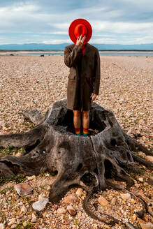 Unrecognizable person in red hat and coat standing in stump center on shore with crushed stones in summer - ADSF31445