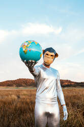 Anonymous guy in silver latex suit with geometric monkey mask looking at camera and giving globe in nature - ADSF31452