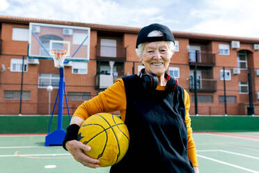 Optimistische reife Frau in Sportkleidung und Kopfhörern, die in die Kamera blickt, während sie auf einem öffentlichen Basketballplatz steht und mit einem Ball trainiert - ADSF31459