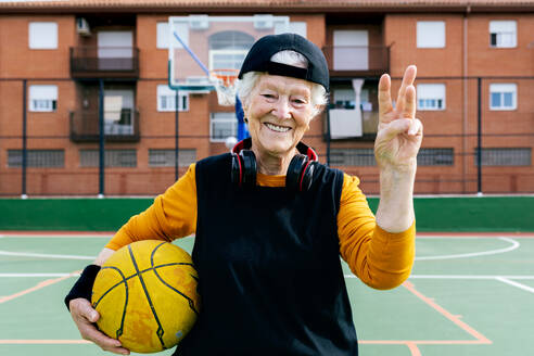 Optimistische reife Frau in Sportkleidung und Kopfhörern, die in die Kamera blickt, während sie auf einem öffentlichen Basketballplatz steht und mit einem Ball trainiert - ADSF31460
