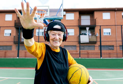 Smiling mature female in headphones with ball in hand looking at camera while standing on sports ground during basketball training - ADSF31462