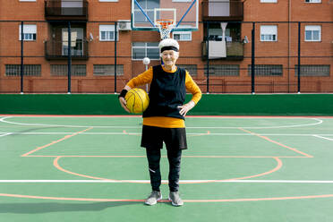 Positive reife Frau in Sportkleidung und Stirnband, die in die Kamera schaut, während sie während eines Basketballspiels mit dem Ball steht - ADSF31466