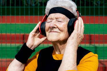 Content mature female with pierced nose in active wear listening to songs in headphones while standing sports ground near net - ADSF31469