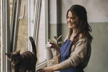 Smiling woman with jar looking at cat in living room - SSGF00223