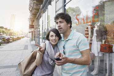 Frau zeigt auf ihren Freund, der mit seinem Smartphone vor einem Geschäft auf dem Gehweg steht - RORF02877