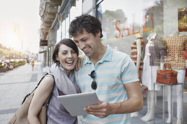 Fröhliches Paar, das ein digitales Tablet benutzt, während es vor einem Bekleidungsgeschäft steht - RORF02879