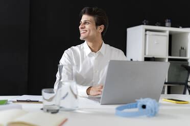 Glücklicher Geschäftsmann sitzt mit Laptop am Schreibtisch im Büro - GIOF14040
