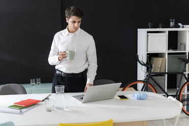 Junger Geschäftsmann mit Kaffeetasse und Laptop im Büro - GIOF14047