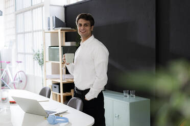 Smiling businessman with hand in pocket having coffee in office - GIOF14048