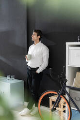 Contemplated businessman with coffee cup leaning on black wall in office - GIOF14049