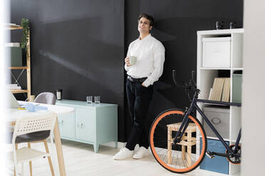 Junger Geschäftsmann mit Kaffeetasse lehnt an schwarzer Wand in kreativem Büro - GIOF14050