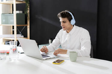 Businessman wearing headphones working on laptop at desk in office - GIOF14052