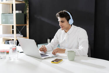 Geschäftsmann mit Kopfhörern bei der Arbeit am Laptop am Schreibtisch im Büro - GIOF14052