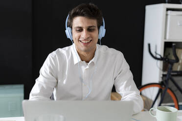 Lächelnder kreativer Geschäftsmann mit Kopfhörern und Laptop im Büro - GIOF14053