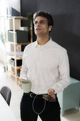 Geschäftsmann mit Kaffeetasse und Smartphone mit Kopfhörern im Büro - GIOF14055