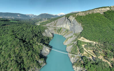 Luftaufnahme einer Brücke über einen See, umgeben von grünem Wald und schneebedeckten Bergen, Monteynard, Lavard, Isere, Rhône-Alpes, Frankreich. - AAEF13293
