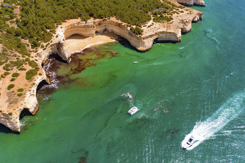 Aerial view of a boat, catamaran and a yacht cruising along the sea near the cliffs of Praia da Corredoura beach in Lagoa, Algarve, Portugal. - AAEF13311