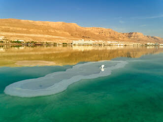 Luftaufnahme eines Plastikstuhls im farbenfrohen Wasser des Toten Meeres und Berge und Hotels am Horizont, Totes Meer, Negev, Israel. - AAEF13314