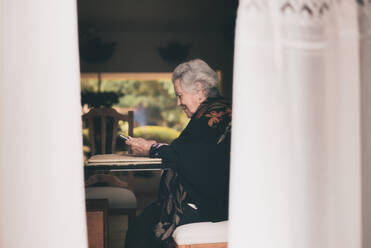 Side view of smiling elderly female wearing warm clothes sitting at table with tablet and cup of tea looking at screen - ADSF31502